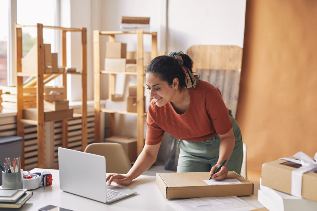 smiling woman typing on laptop and accepting the orders then packing parcels for shipment