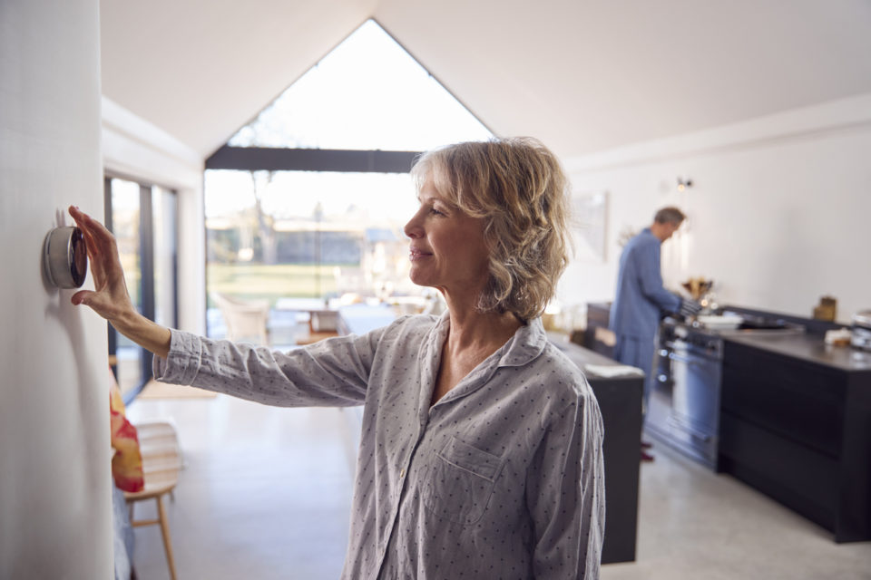 Mature Woman Adjusting Wall Mounted Digital Central Heating Thermostat Control At Home